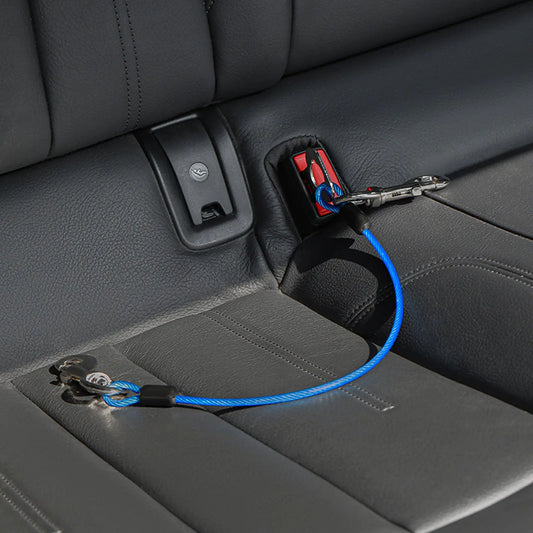 A blue braided car seatbelt lock and release device installed on a gray leather car seat, with the lock attached to a seatbelt clip.